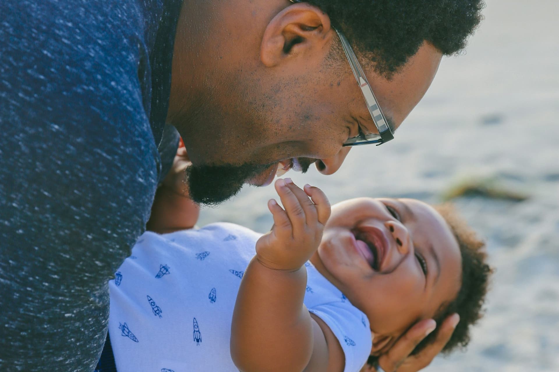 A smiling father in glasses holds his laughing baby close on a sandy beach